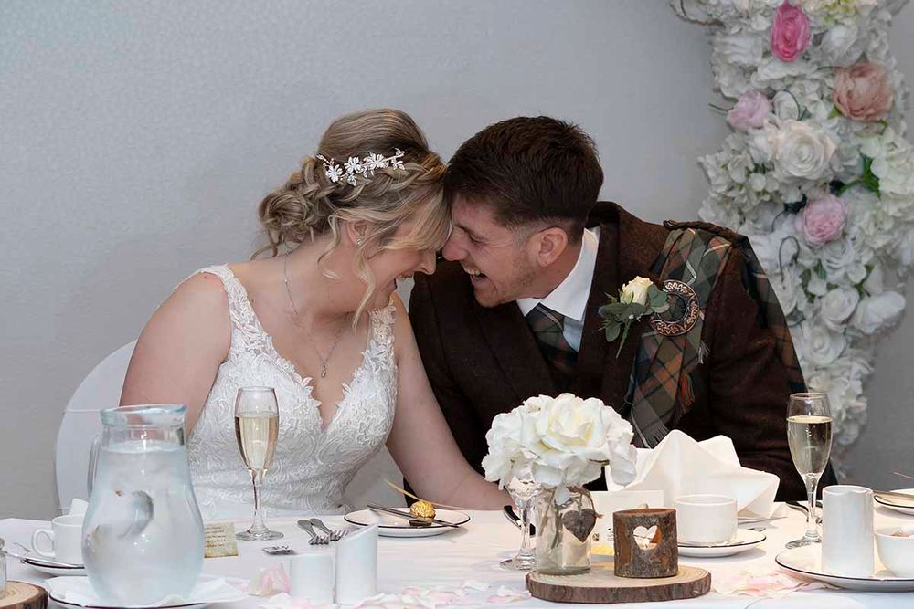 Bride and groom share an intimate moment at wedding reception table with champagne glasses and floral decorations.