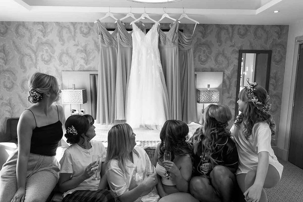 Black and white photo of bridesmaids admiring a wedding dress hanging in a hotel room.