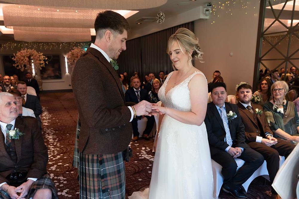 Groom in tartan kilt places wedding ring on bride in white gown during indoor ceremony with seated guests.