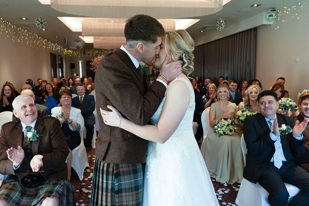 Bride and groom share first kiss at Scottish wedding ceremony with guests applauding in background.