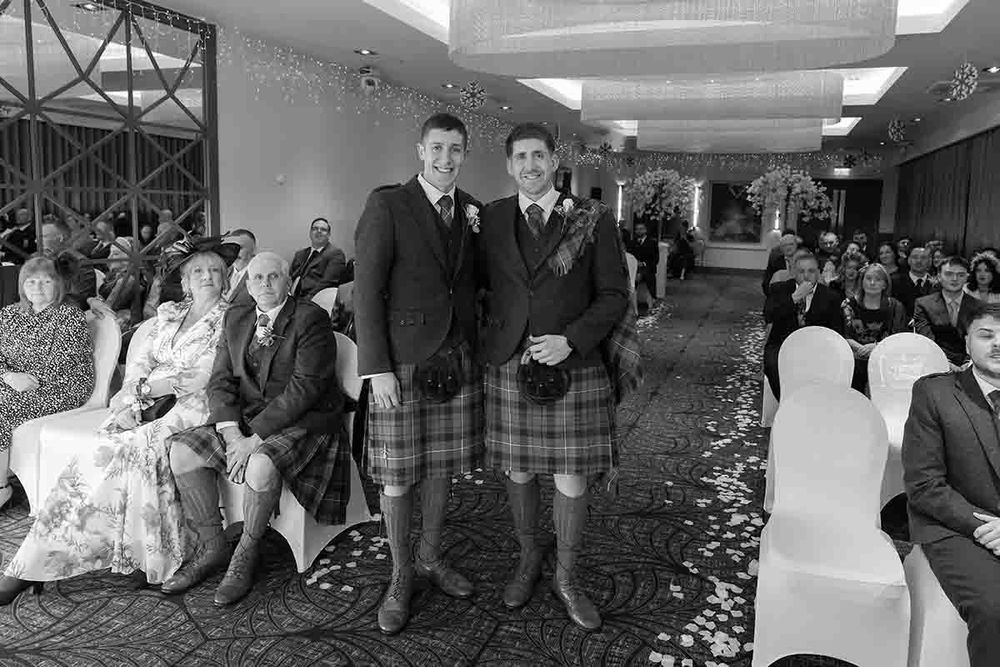 Two grooms in Scottish kilts stand together at their wedding ceremony in a decorated venue with seated guests.