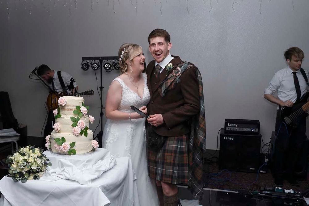 Bride and groom laughing together while cutting their wedding cake, groom wearing traditional Scottish tartan kilt.