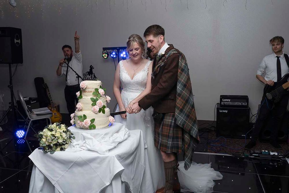Bride and groom cutting their wedding cake together, groom wearing traditional Scottish tartan kilt, live band performing in background.