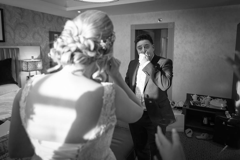 Black and white photo of groom covering his mouth in awe as bride reveals wedding dress during emotional first look.
