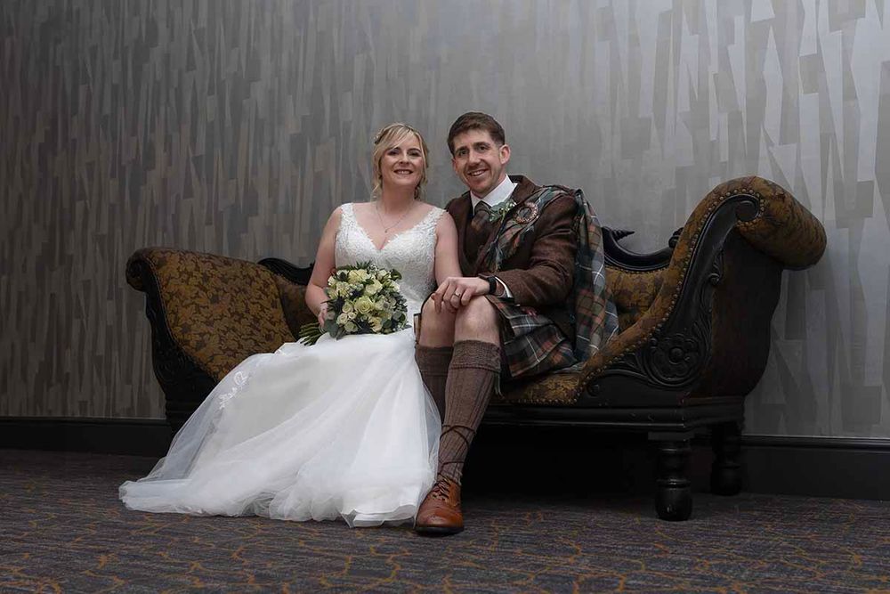 Bride in white gown and groom in Scottish tartan kilt sit together on ornate chaise lounge for wedding portrait.