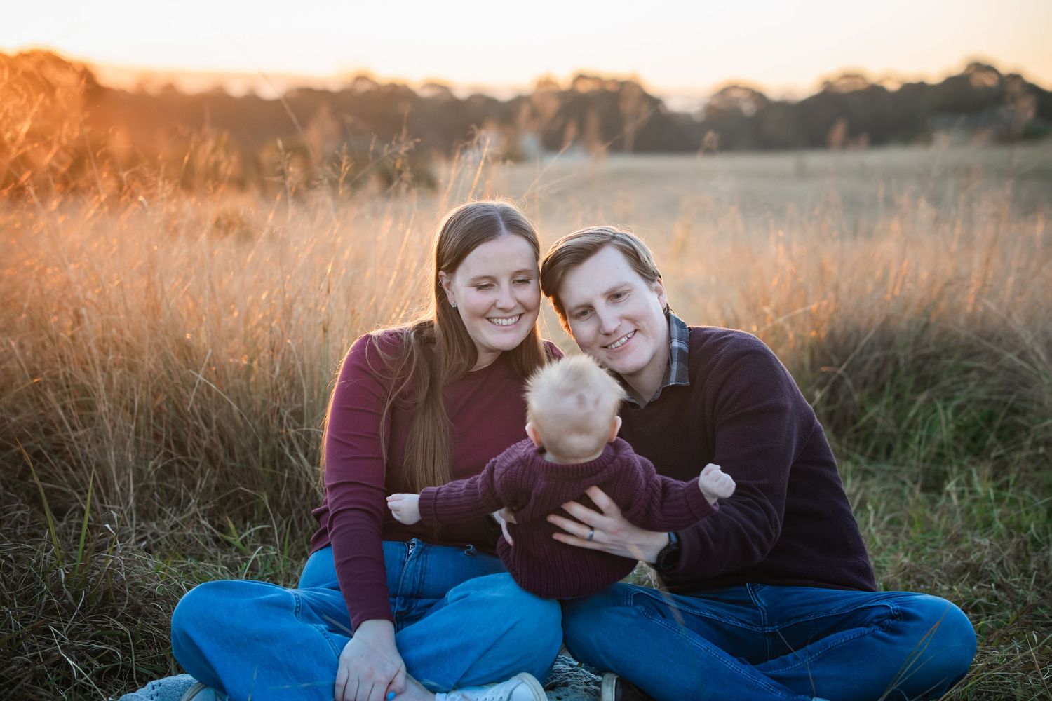 Happy family with baby sitting in golden field at sunset, smiling couple at a family photoshoot at Rouse Hill Regional Park