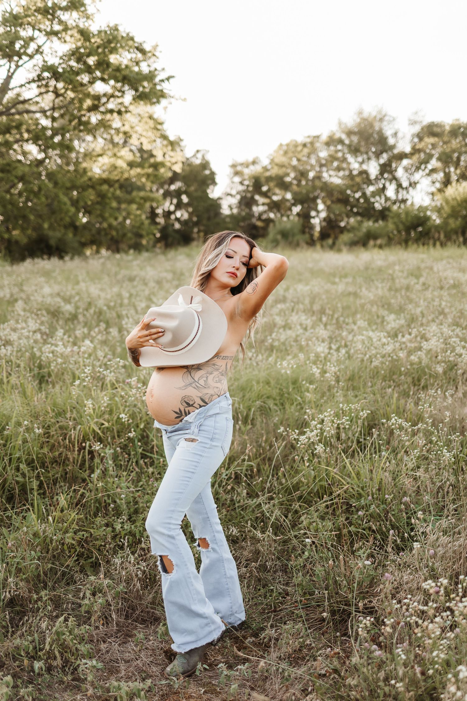 Pregnant woman in ripped jeans holding cowboy hat in a wildflower field at golden hour.
