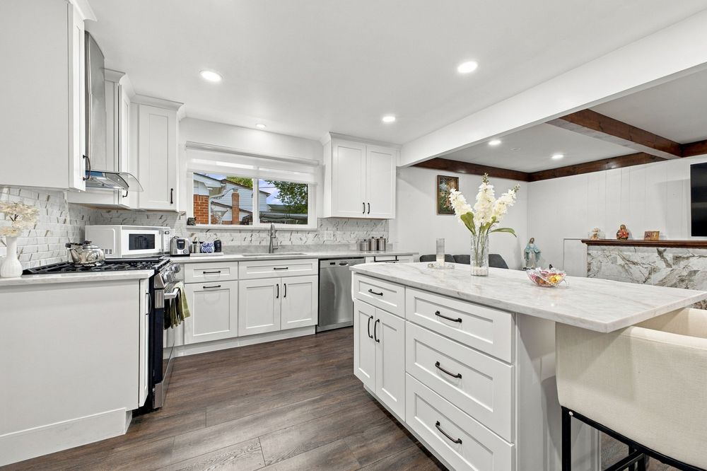 Modern white kitchen with marble island, shaker cabinets, stainless appliances, herringbone backsplash, and hardwood floors.