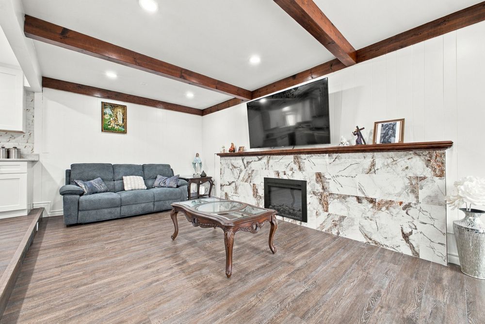 Living room with beamed ceiling, hardwood floors, marble fireplace, wall-mounted TV, and gray sofa with coffee table.