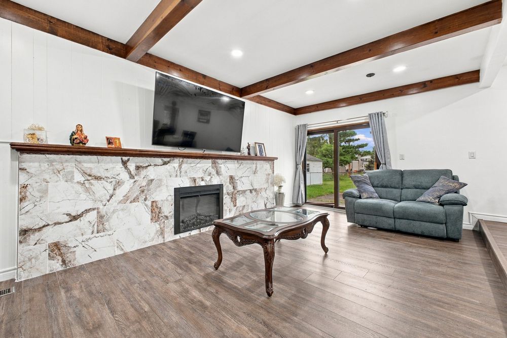 Living room with marble fireplace, wood beam ceiling, hardwood floors, wall-mounted TV, and recliner sofa near sliding glass door.