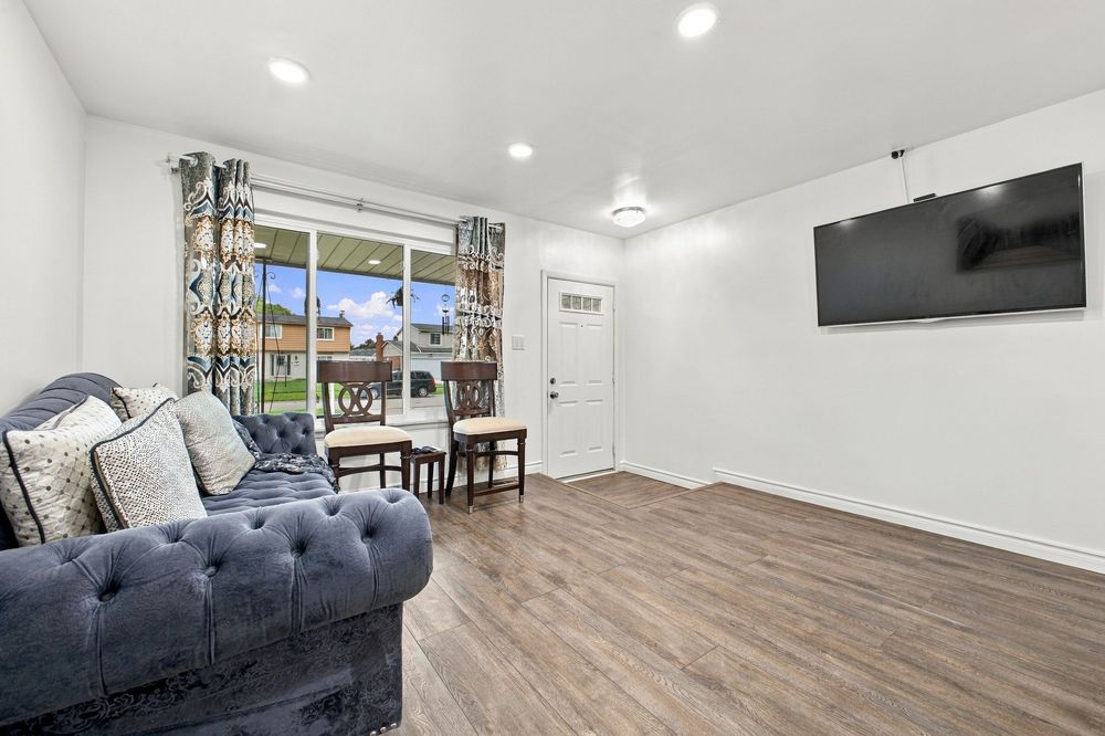 Bright living room with gray tufted sofa, hardwood floors, wall-mounted TV, and large window overlooking a yard.
