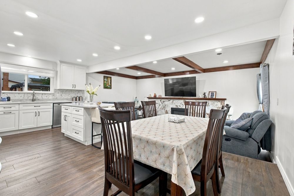 Open-concept dining area with beamed ceiling, dark hardwood floors, white kitchen cabinets, and cozy living space in background.