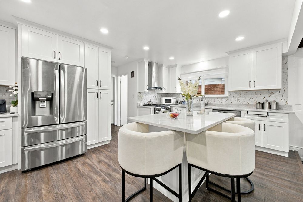 Modern white kitchen with stainless steel fridge, island seating, quartz countertops, and dark hardwood floors.