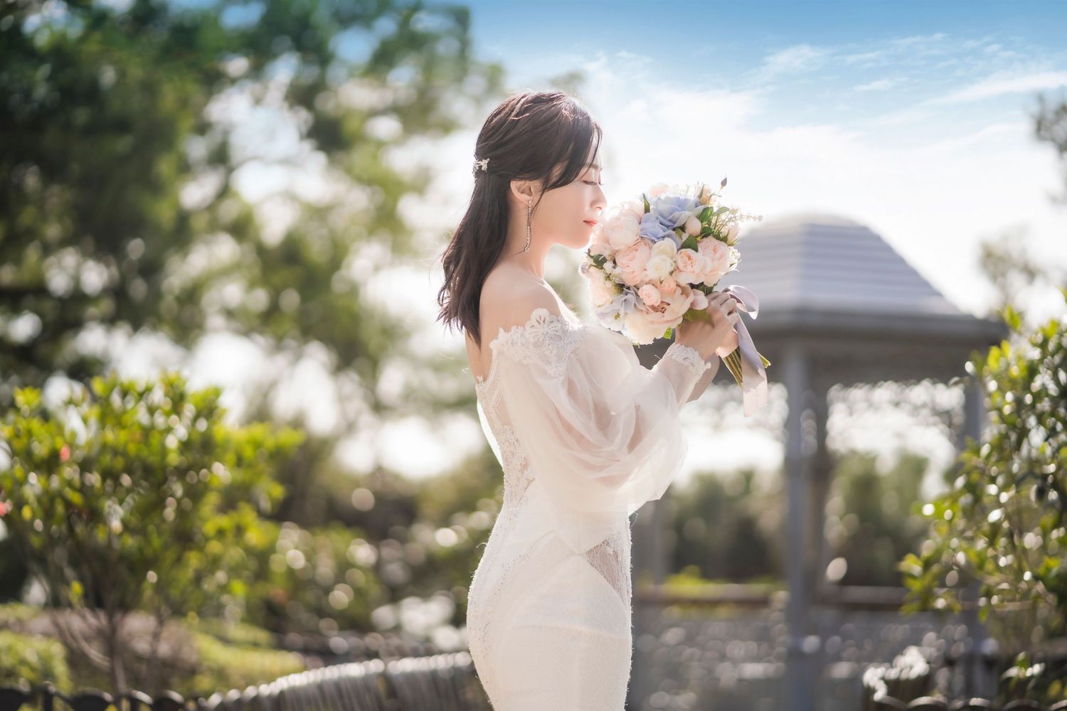 Elegant bride in white lace dress holds pastel bouquet while posing in sunlit garden with gazebo in background.