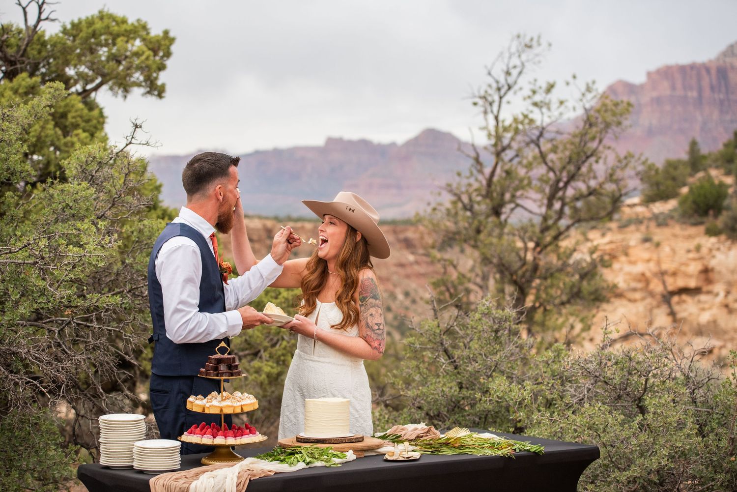 Couple exchanging vows outdoors with officiant, red rock canyon backdrop, bride in lace gown, groom in navy vest.