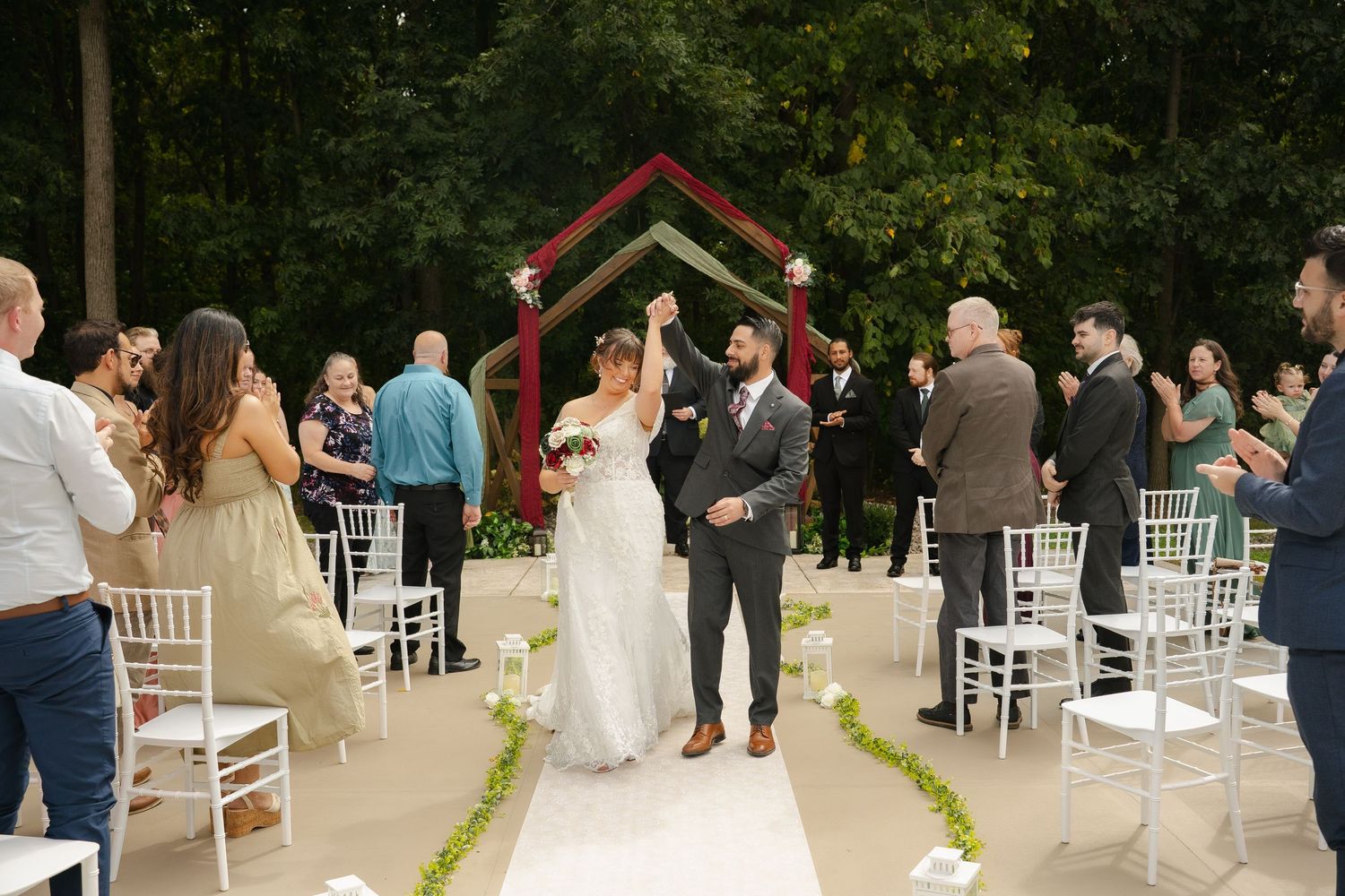 Newlywed couple raising hands at outdoor wedding ceremony with green arch backdrop and white chairs.