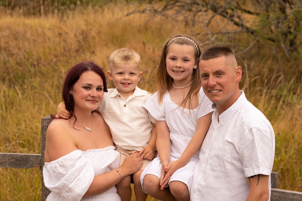 Happy family of four in white outfits posing outdoors in a golden field near a wooden fence.