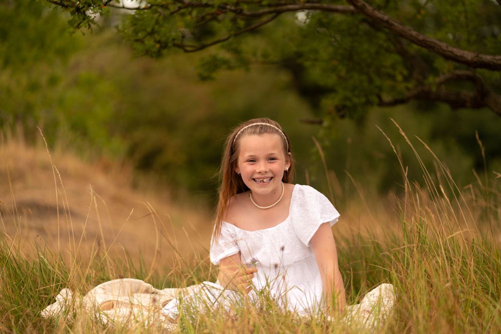 A smiling young girl in a white dress sits in tall golden grass outdoors, surrounded by lush green trees.