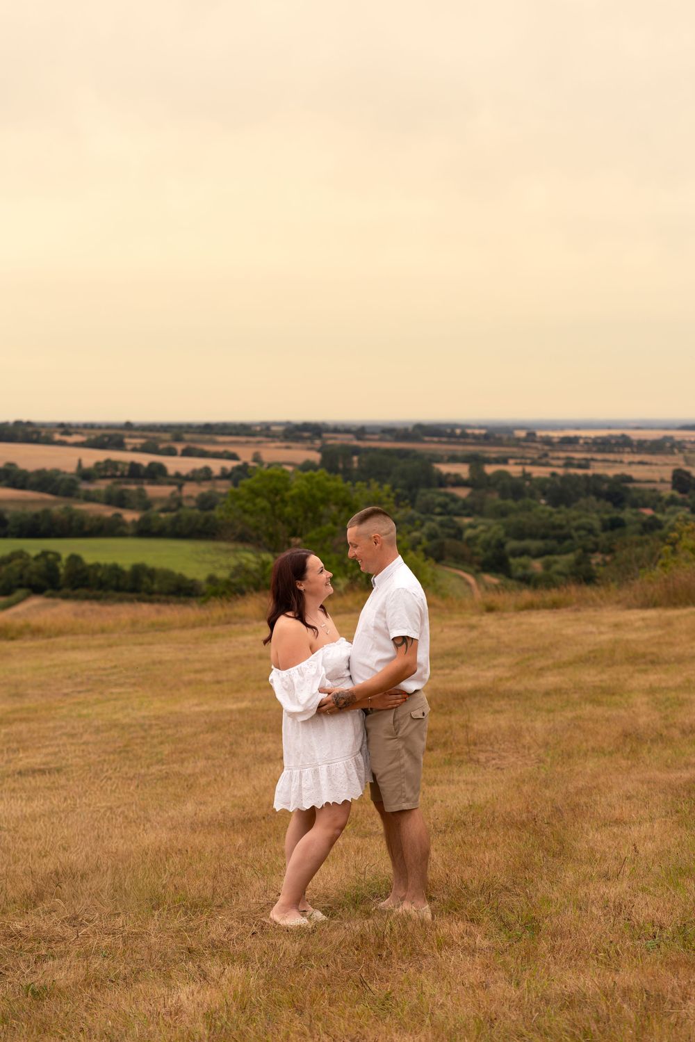 Couple in white outfits embracing on a grassy hill with scenic countryside fields in the background.