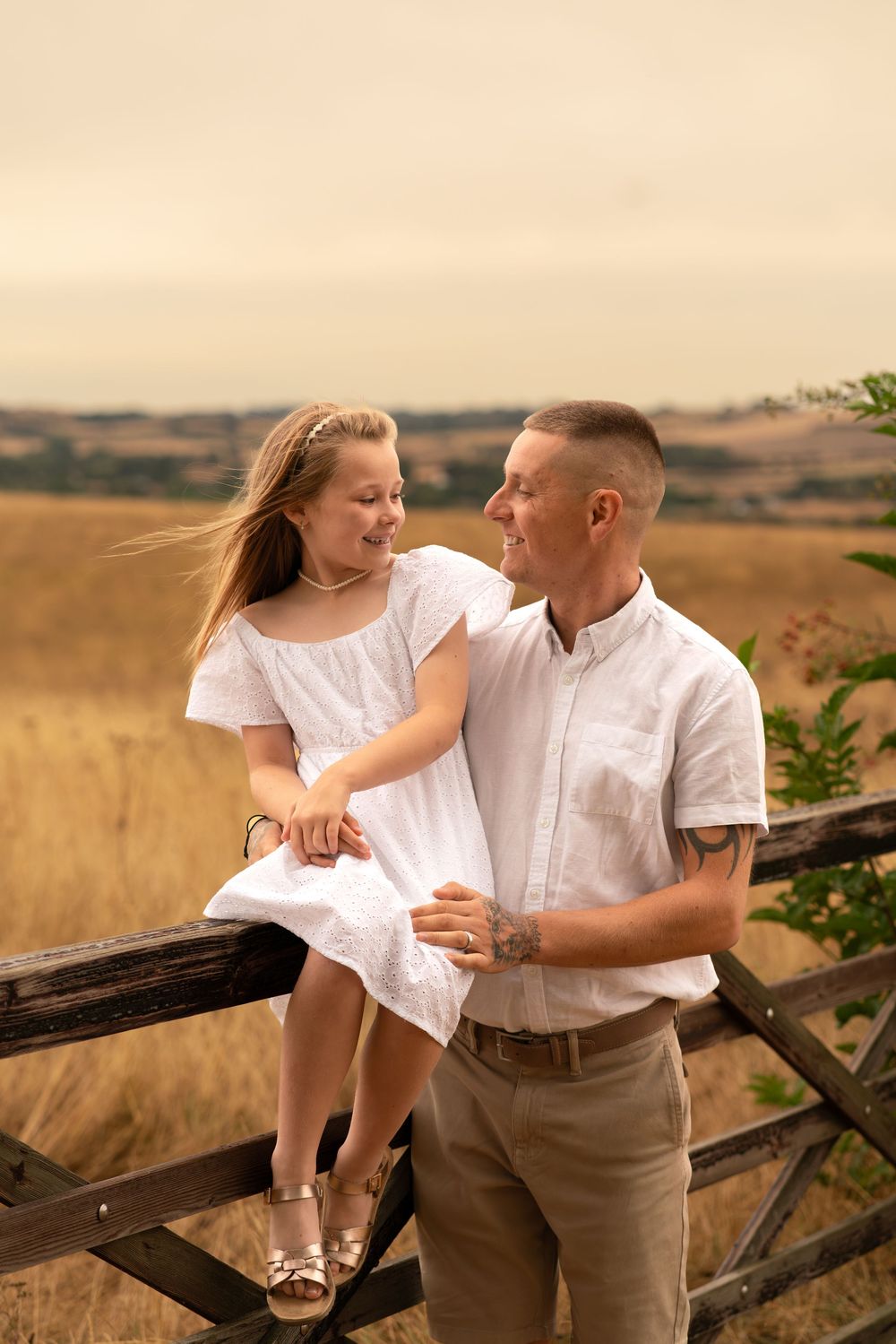 A couple in white and beige outfits share a smile near a rustic wooden fence in a golden field.
