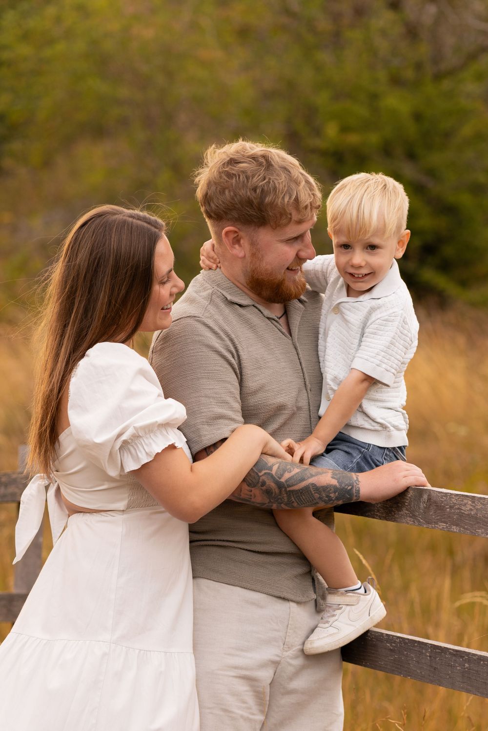 A happy young family with a toddler boy sitting on a wooden fence in a sunny autumn field.