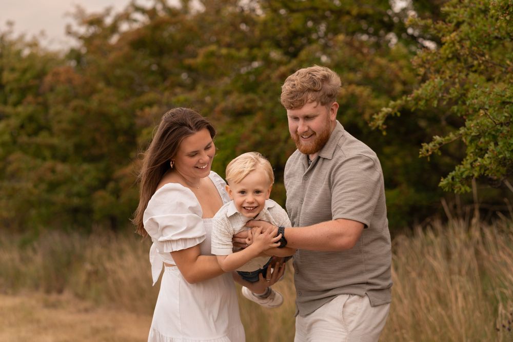 A happy young family with a toddler boy poses outdoors in a golden field with autumn trees in the background.