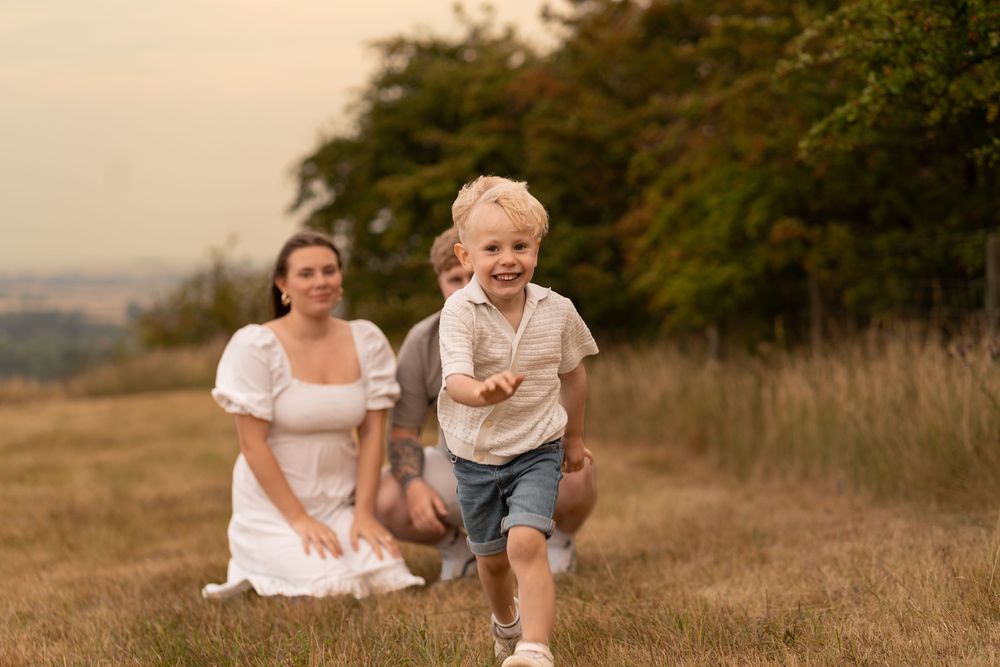 A joyful blonde toddler runs through a grassy field while his mother in a white dress watches warmly at sunset.