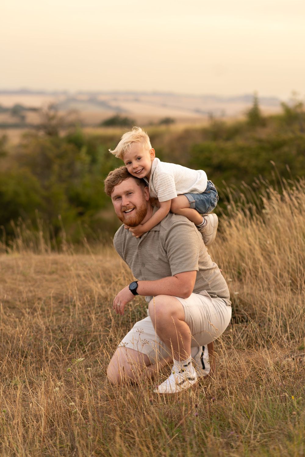 A bearded father crouches in a field while his young blonde child rides on his back, smiling outdoors.
