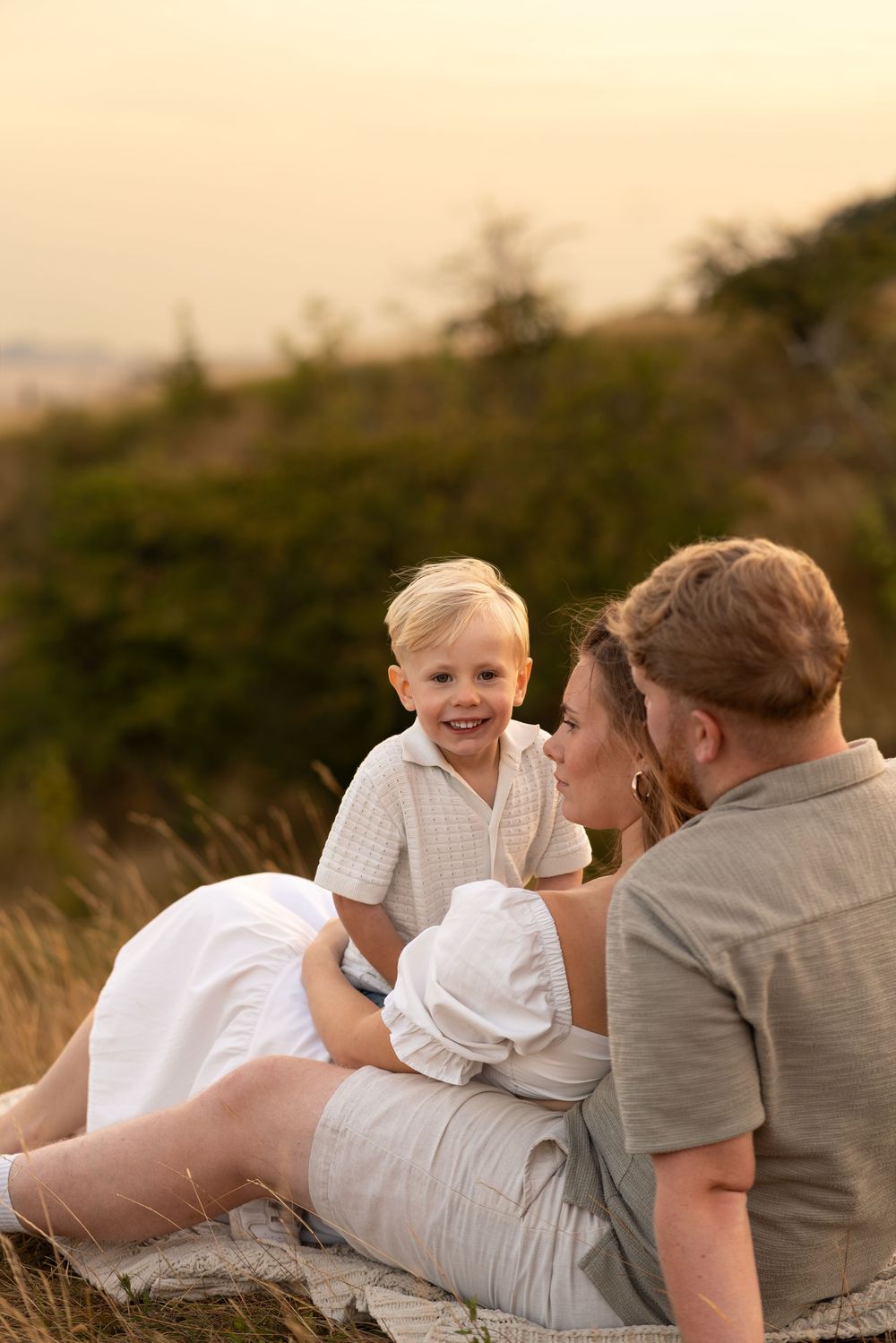 Family relaxing outdoors at sunset, with a smiling toddler boy sitting with his parents in a golden field.