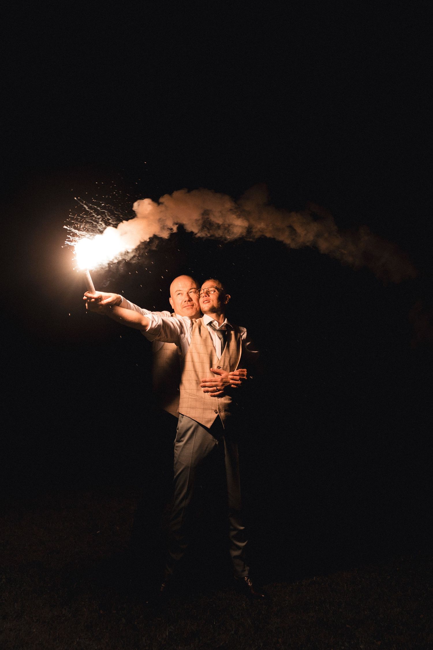 Couple embracing at night holding a bright sparkler with smoke trails against a dark sky backdrop.