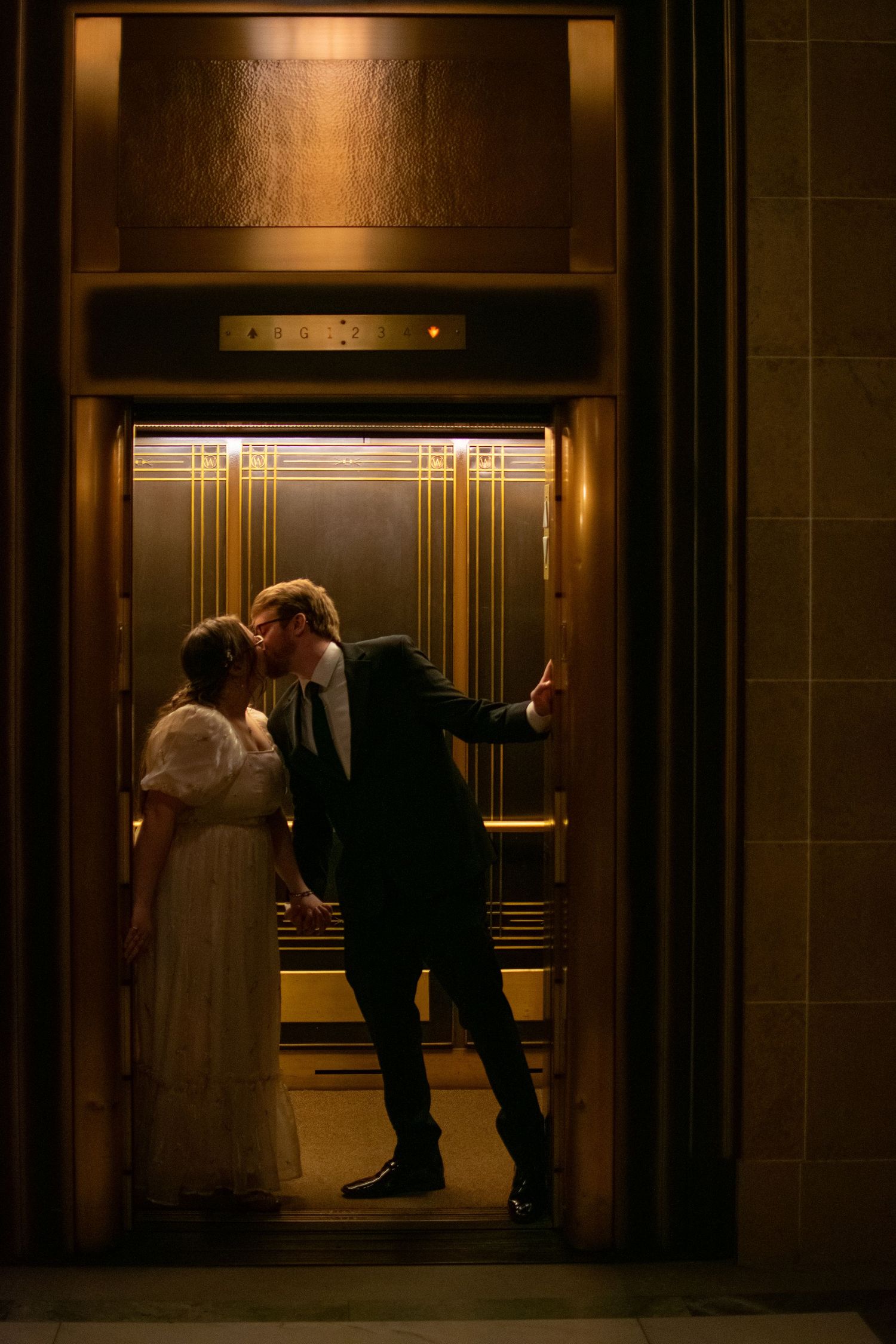 Bride and groom sharing a romantic kiss inside a vintage gold Art Deco elevator, dressed in wedding attire.