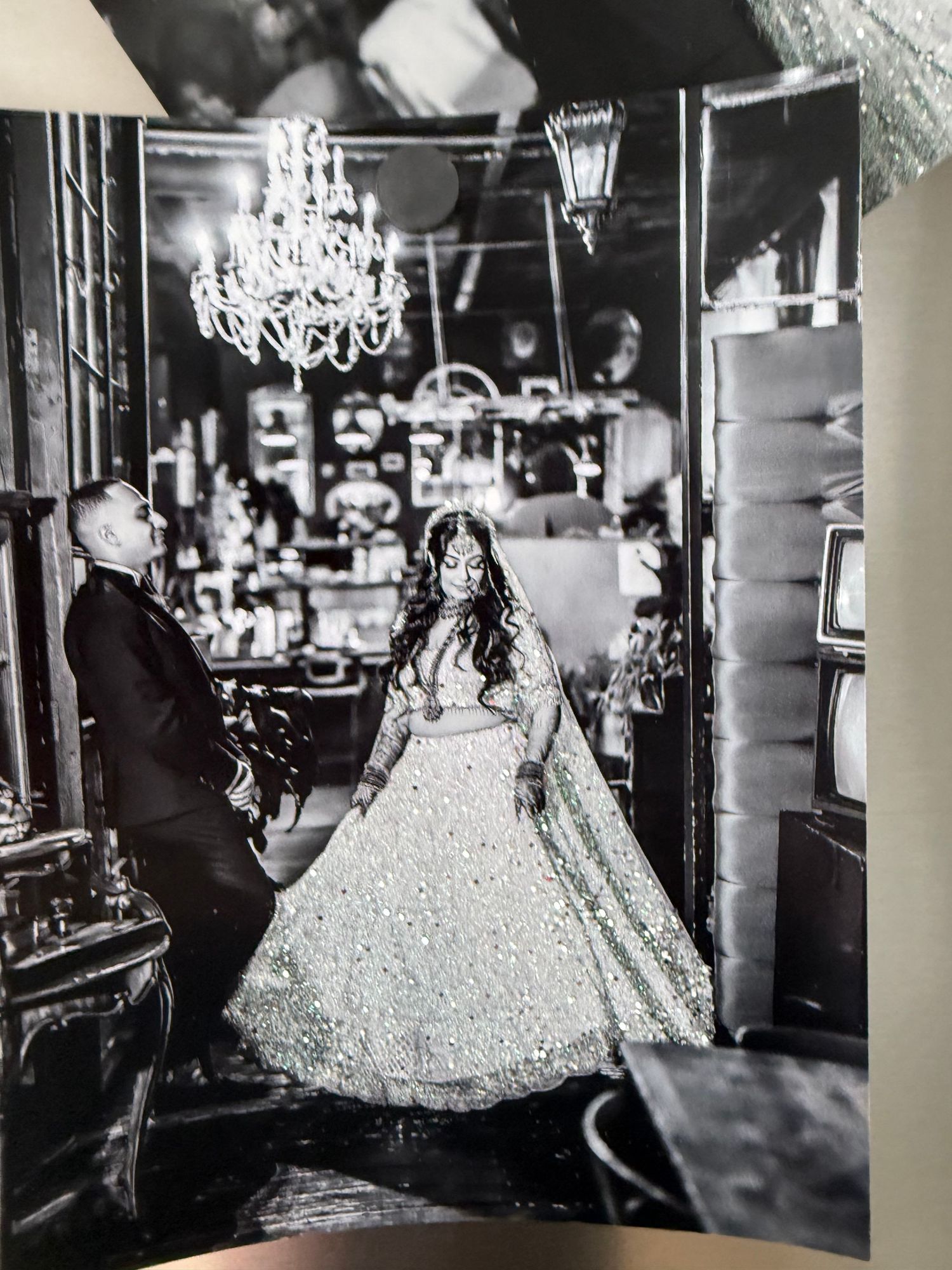 Black and white photo of a bride in an ornate ballgown standing in an elegant venue with a chandelier overhead.