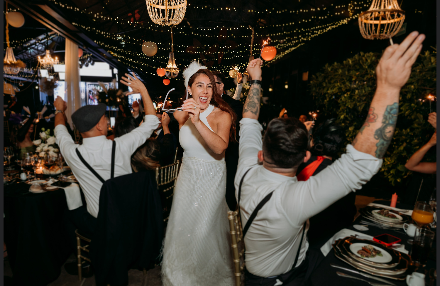 Bride in white dress dancing joyfully with guests at an elegant outdoor wedding reception with string lights.