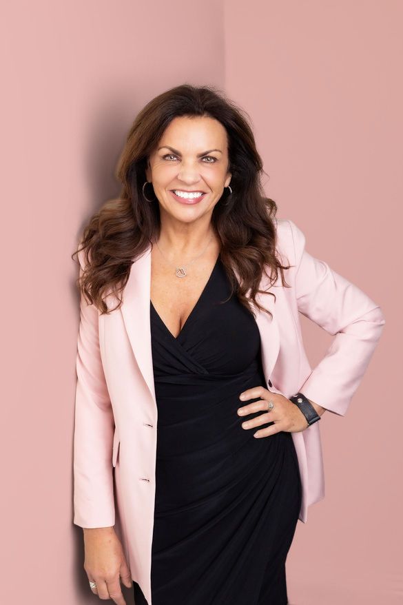 Smiling woman in pink blazer and black dress poses confidently against a pink wall, wearing hoop earrings and a smartwatch.