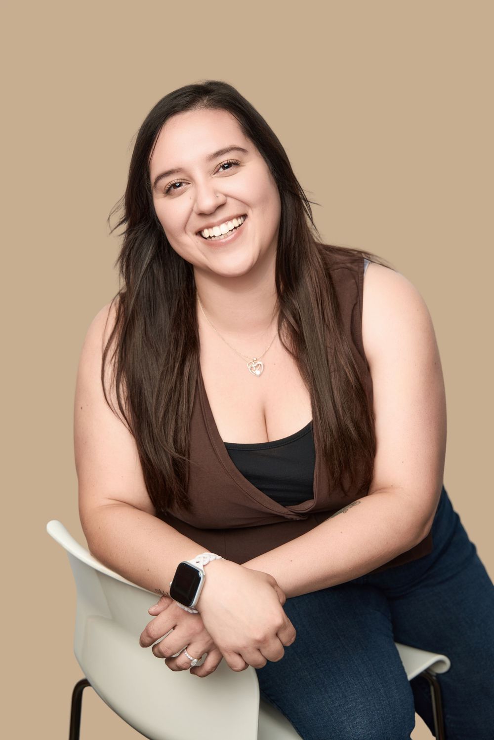 Smiling young woman with long brown hair sitting on a chair, wearing a brown top and jeans against a beige background.