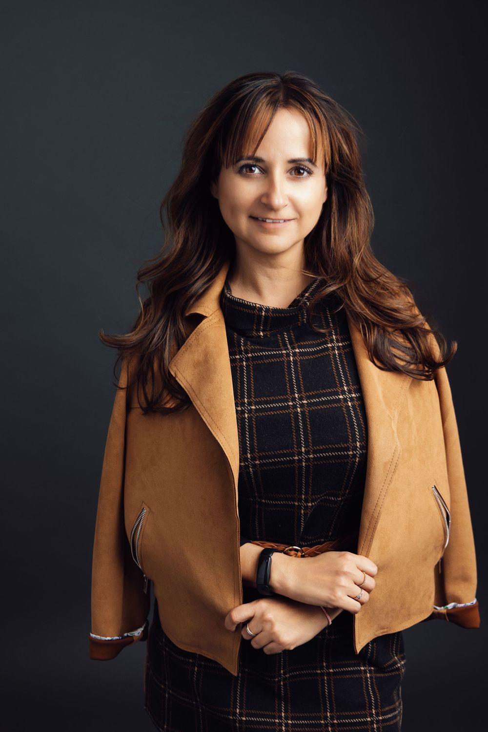 Smiling woman with brown hair wearing a plaid dress and tan suede jacket poses confidently against a dark background.