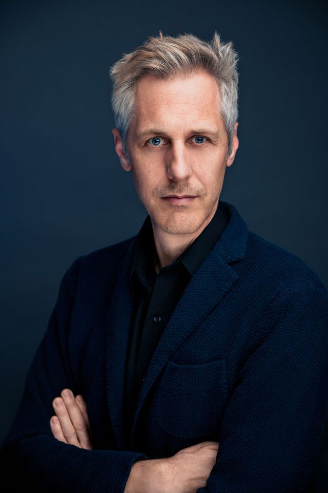 Professional headshot of a middle-aged man with silver hair, blue eyes, arms crossed, wearing a navy blazer against dark background.