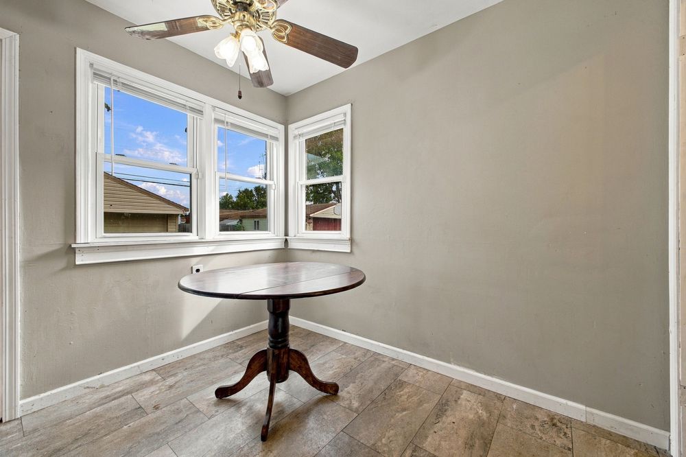 Bright dining nook with round pedestal table, ceiling fan, hardwood floors, and two windows overlooking neighborhood.