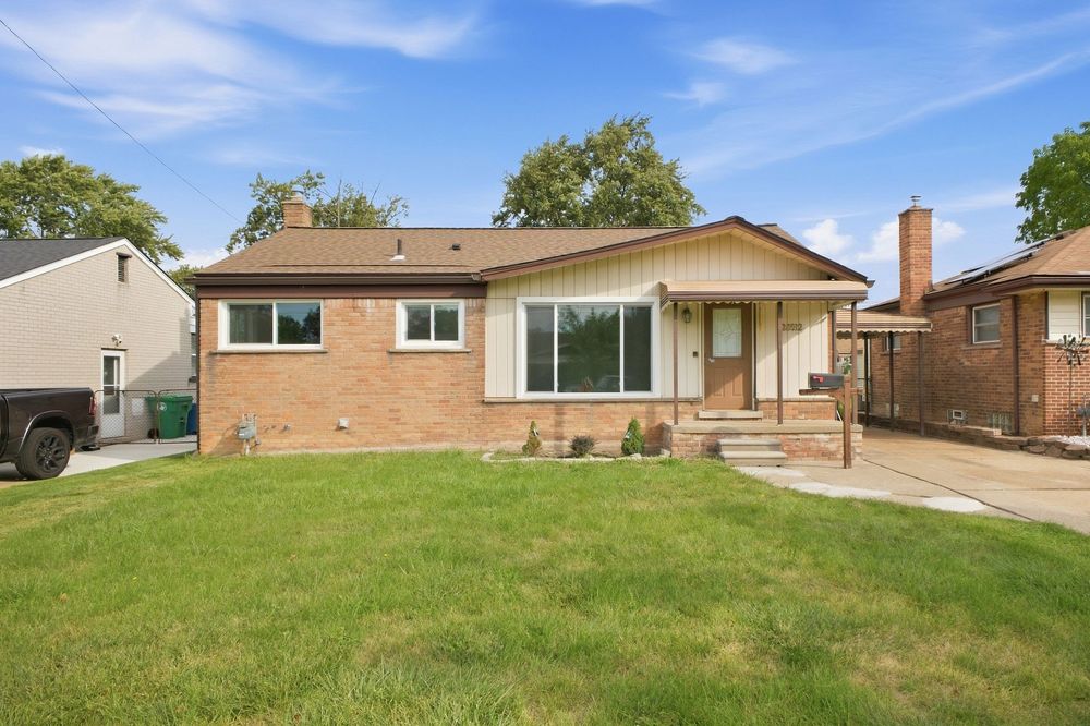 Brick ranch-style home with green lawn, covered porch, and mature trees under a blue sky.