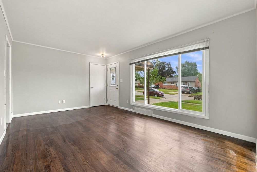 Bright empty living room with dark hardwood floors, crown molding, chandelier, and large window overlooking a suburban street.