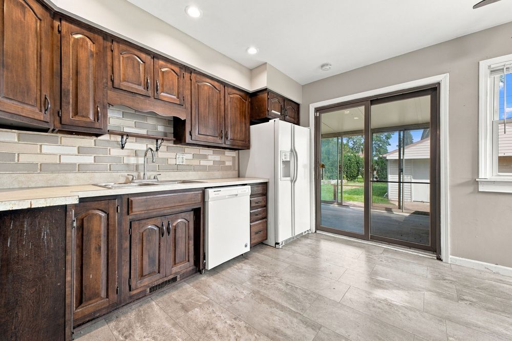 Modern kitchen with dark wood cabinets, tile backsplash, white appliances, and sliding glass doors leading to a bright outdoor patio.