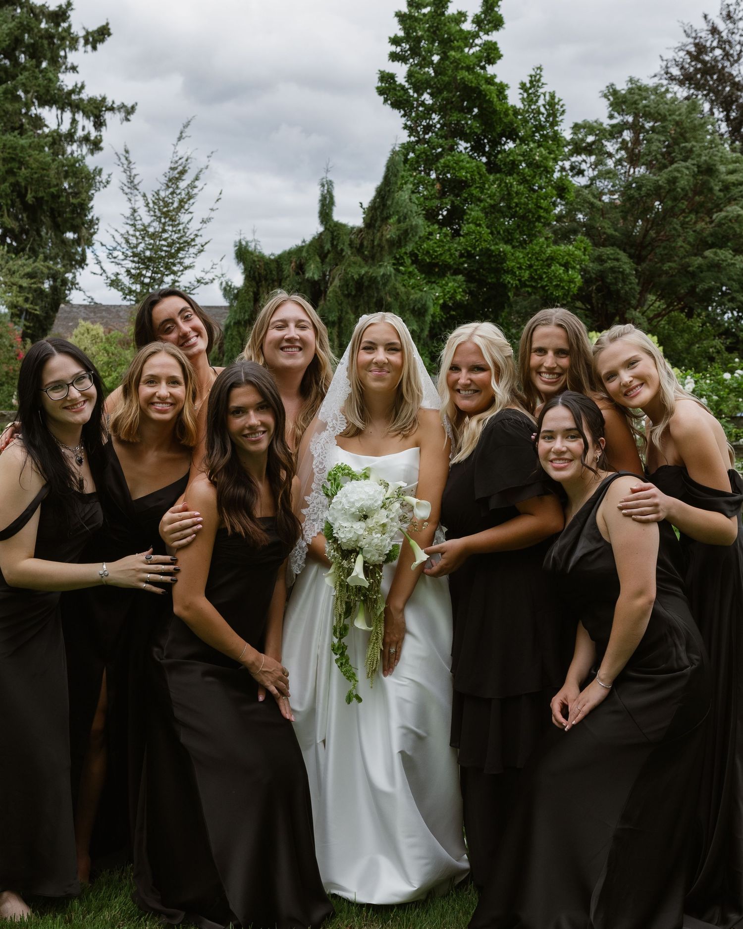 Bride in white gown hugging bridesmaids in black dresses from behind at outdoor wedding ceremony.
