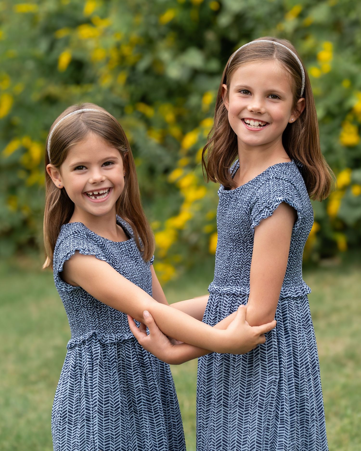 Two young girls in matching blue patterned dresses hold hands and smile outdoors with yellow flowers behind them.