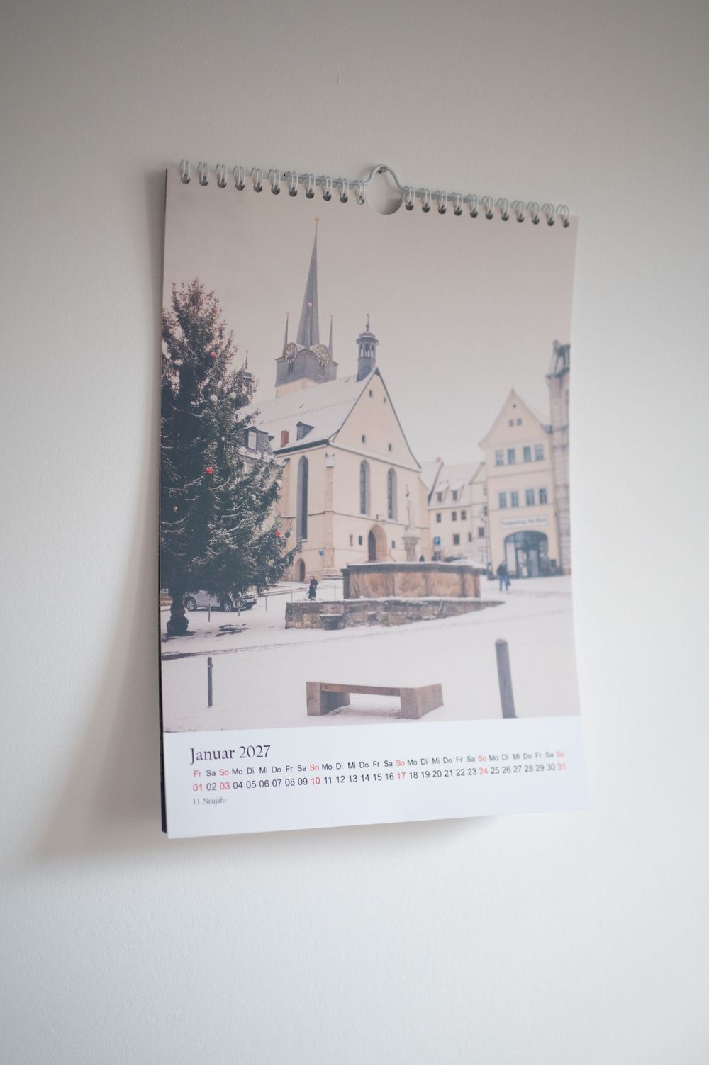 A spiral-bound wall calendar showing Januar 2027 with a winter photo of a European town square with a church.