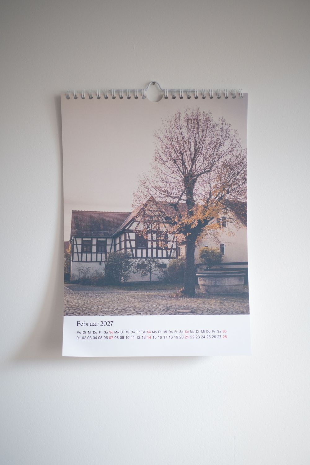 A wall calendar showing February 2027 featuring a photo of a traditional half-timbered house with a bare tree in autumn.