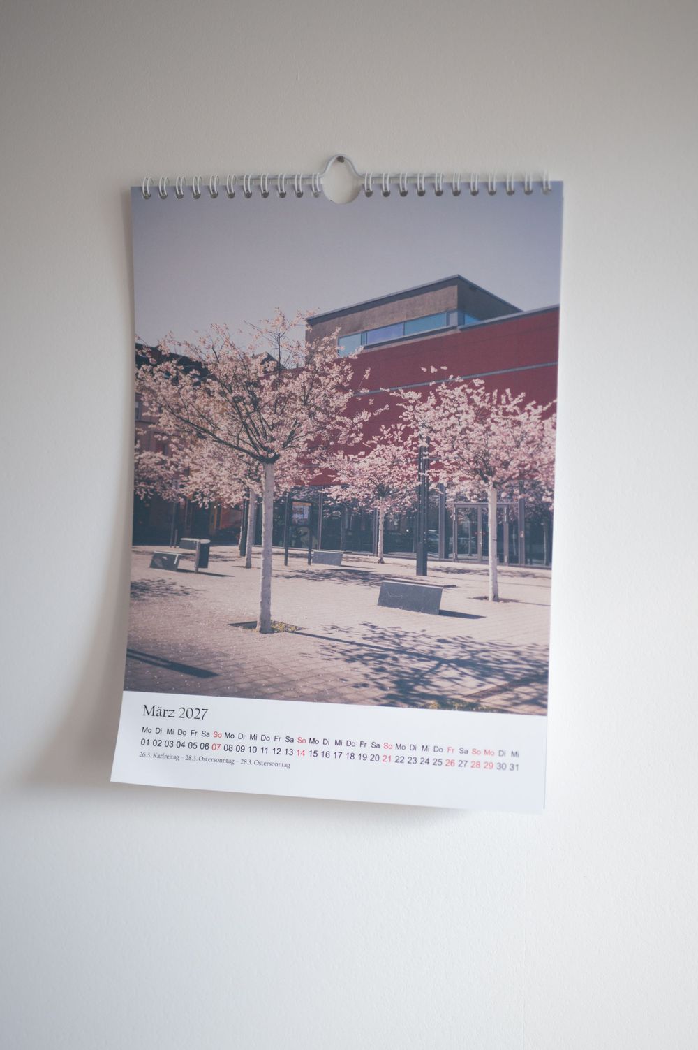 A wall calendar showing März 2027 with a photo of cherry blossom trees in front of a modern red brick building.