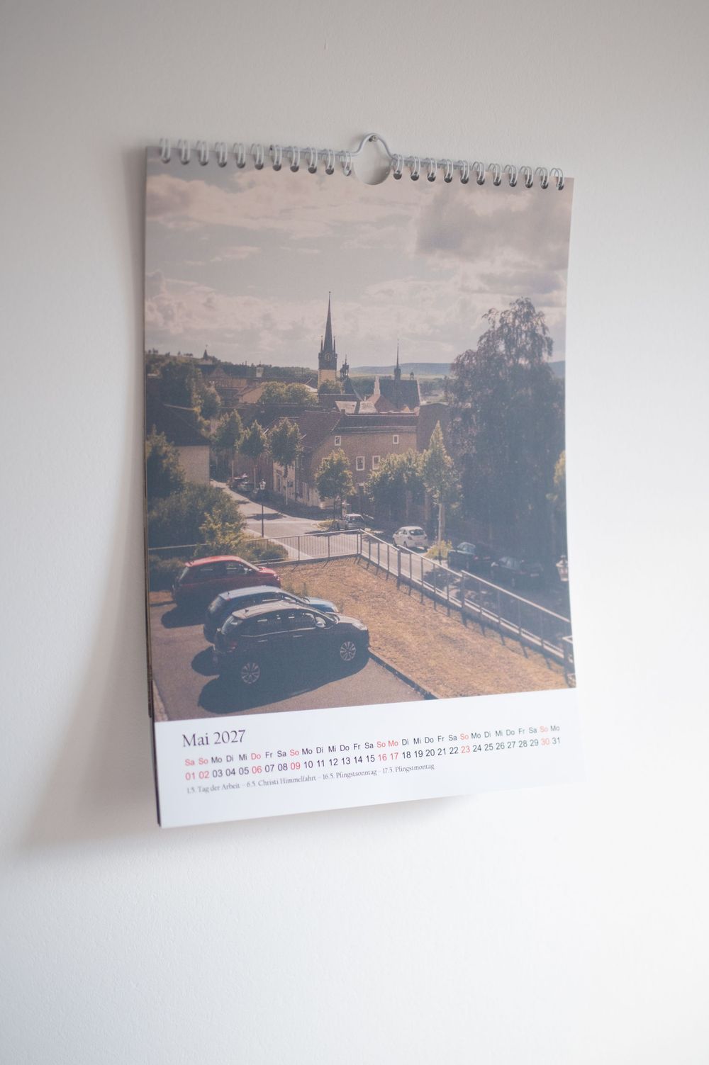 A wall calendar showing May 2027 with a vintage cityscape photo featuring classic cars parked near a church spire.