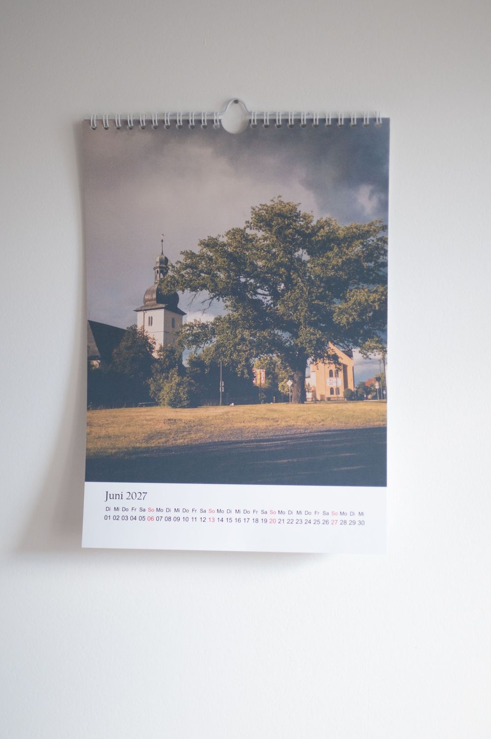 A wall calendar showing Juni 2027 with a scenic photo of a church and large tree under dramatic cloudy skies.