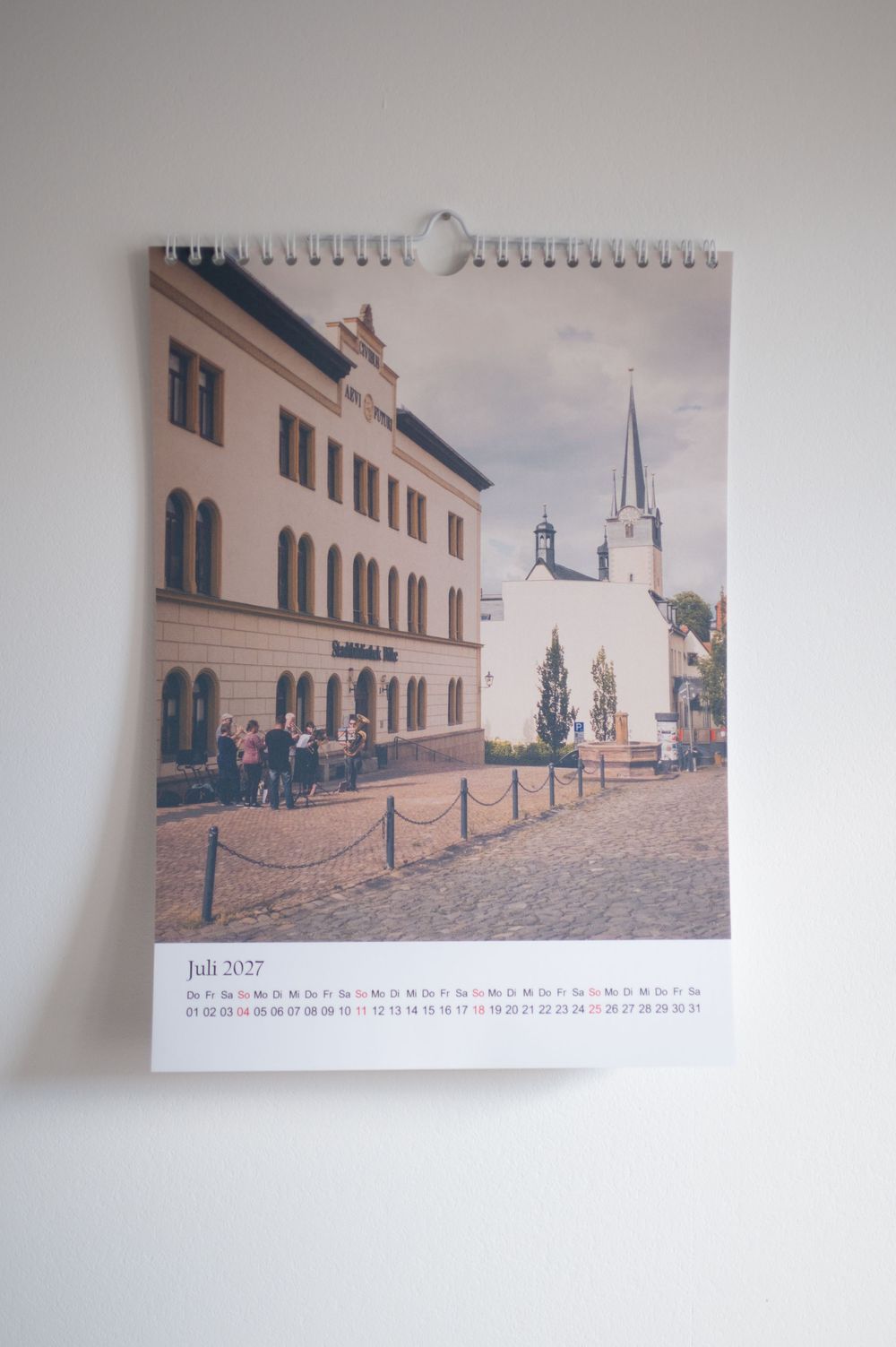 A wall calendar showing Juli 2027 with a photo of a European cobblestone street featuring a church steeple and historic buildings.
