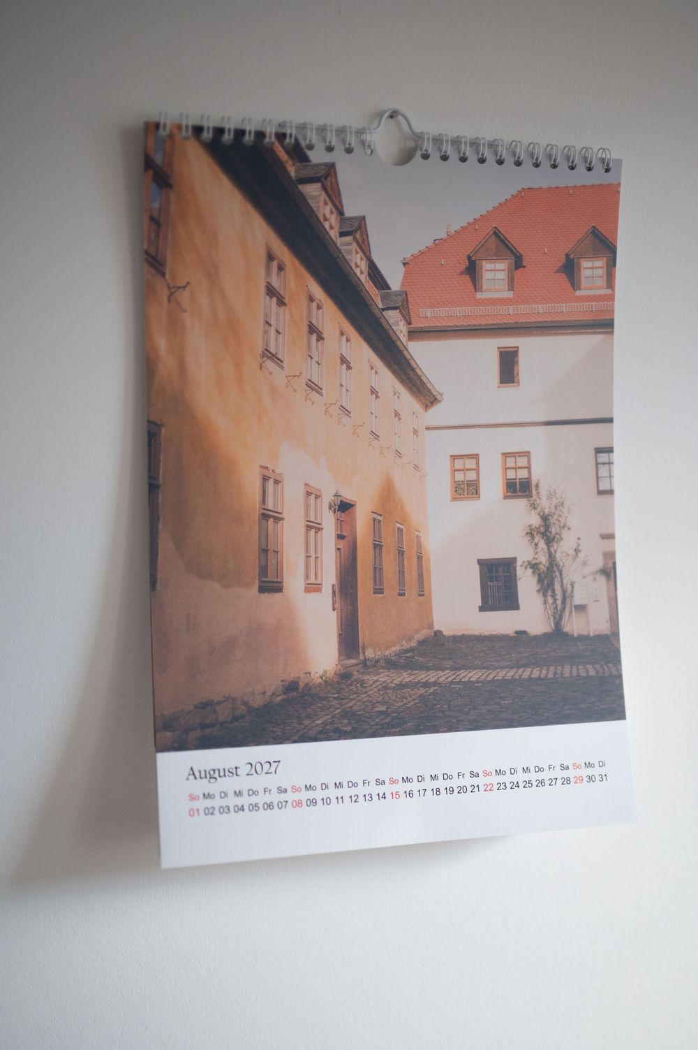 Wall calendar showing August 2027 with a photo of a historic European courtyard featuring orange and white buildings.